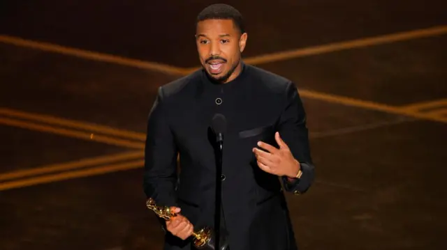 Michael B Jordan wears a black collarless suit and speaks into the microphone while holding his trophy.