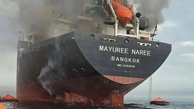 Smoke billows from the black and red cargo ship "Mayuree Naree" as it sits in calm water.