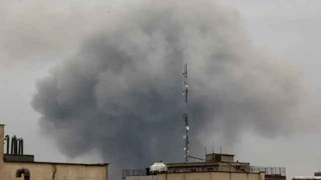 Smoke rises in the shape of a dark grey mushroom cloud following from behind a building