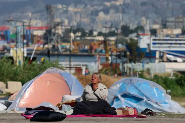 A woman sits on a blanket on a street, tents are proped up behind her and the city scape of Beirut can be seen in the background
