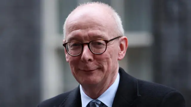 British Secretary of State for Work and Pensions Pat McFadden smiling in a close-up at the camera with a blurry Downing Street behind him