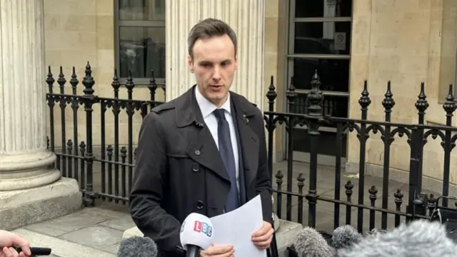 Andrew speaks to reporters outside the court. The building is built of light stone, and has large columns and iron fencing at the front. Andrew has a serious expression and is holding a piece of paper, as lots of fluffy grey microphones point towards him. He has short brown hair and is wearing a black jacket, a white shirt and a navy tie.