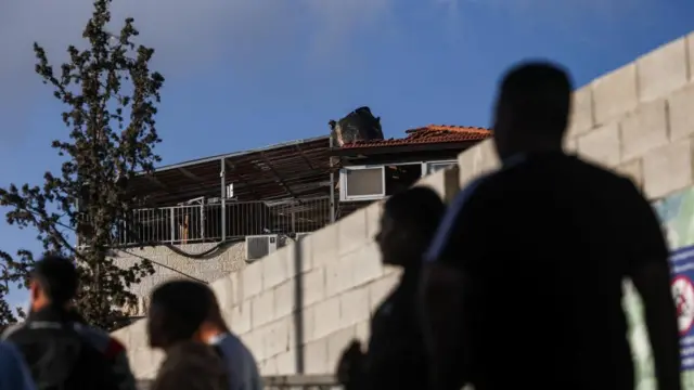 Four people in front of a white wall. On the other side of the wall, part of a missile is visible slightly poking out of the roof of a h