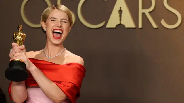 Jessie Buckley, who wears a red and pink gown, jubilantly holds her Oscar with hold hands while standing in front of the word 'Oscars' on a brown wall.