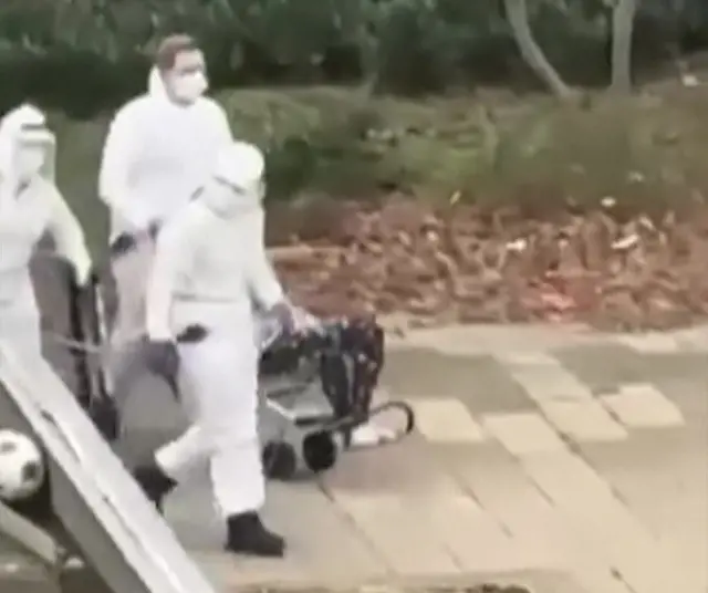 A grab from video shows people in white hazmat suits wheeling a patient down a pavement