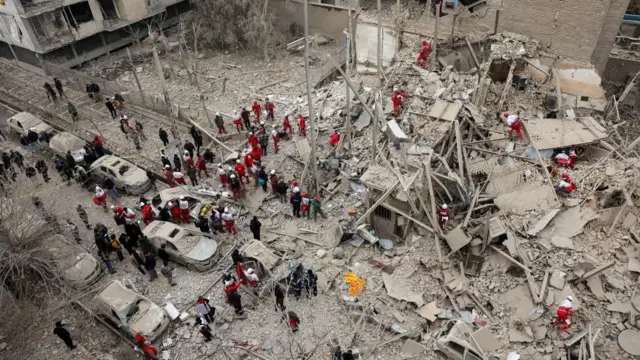 Emergency personnel work at the site of a strike on a residential building. The photo is taken from above and lots of people in black and red and white uniforms are standing next to shattered cars covered in dust and a gutted building