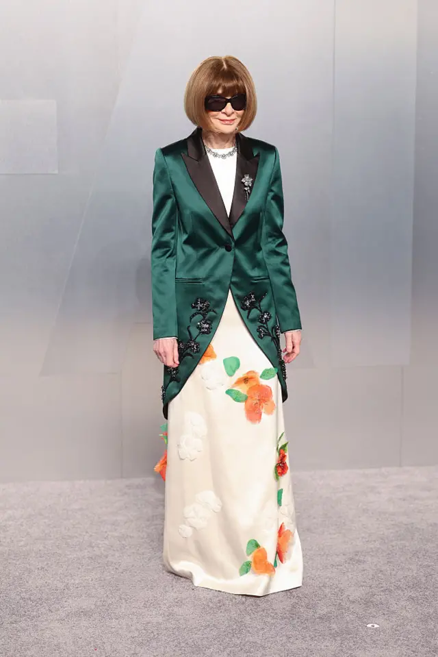 Anna Wintour in a cream dress with orange flowers and a long, green jacket and sunglasses