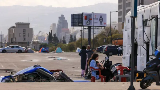 Tents in a car park can be seen with the backdrop of Beirut behind it. A woman sits in a chair on the side of the road with a baby in her lap