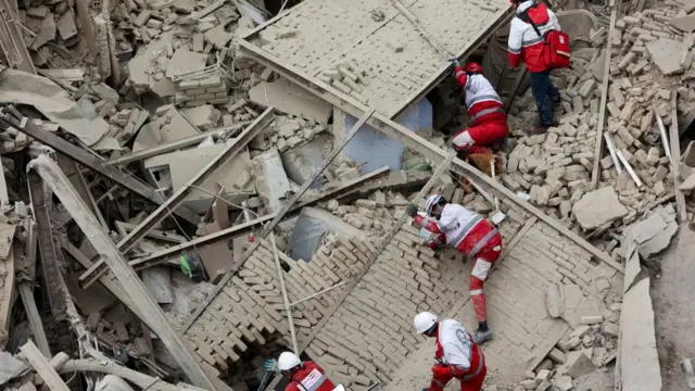 Five emergency workers in red and white uniforms climb through a huge pile of rubble from a building that has been destroyed in Tehran