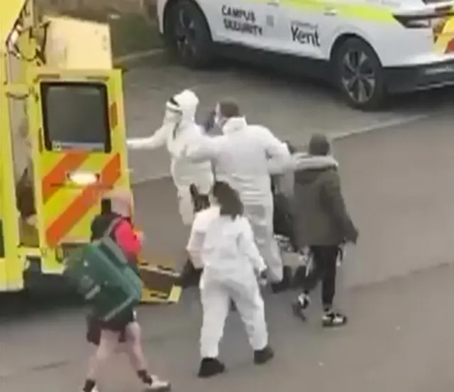 A grab from video shows people in hazmat suits loading a patient into an ambulance with another emergency vehicle saying 'Kent campus security' in the background.