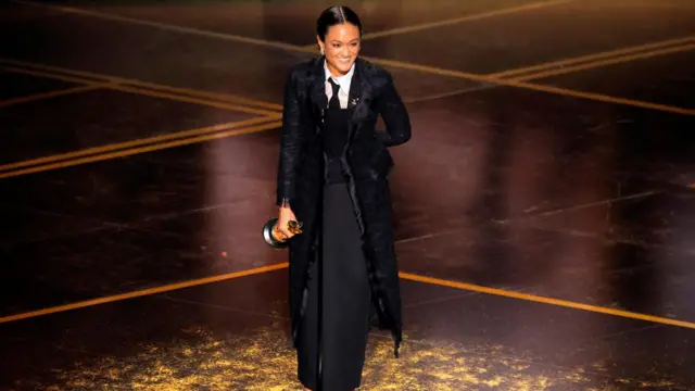 Autumn Durald Arkapaw, dressed in a long black coat over a dark skirt and top, holds her trophy.
