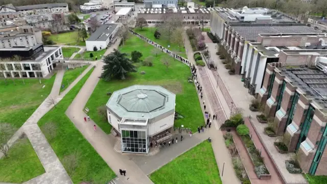 An aerial shot of students queuing - they stand in a long line outside that bends around part of the campus