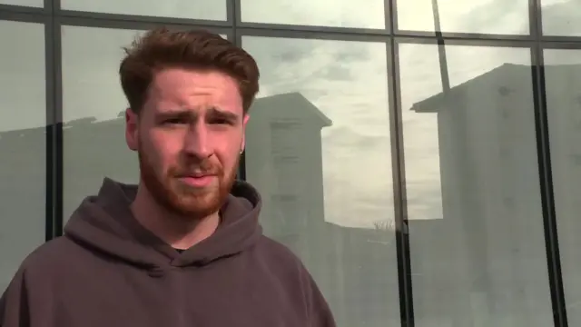 A young man in a hoodie is standing against a building.