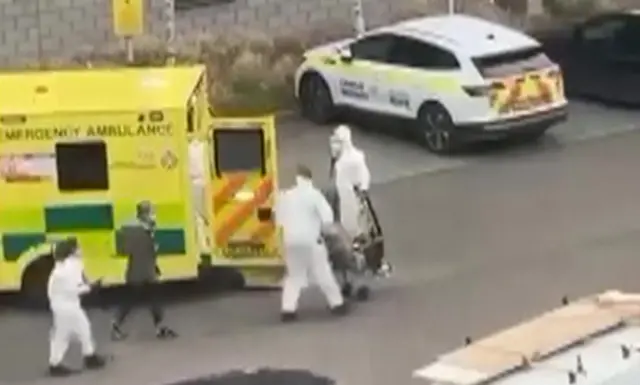 A grab from video shows people in hazmat suits loading a patient into an ambulance with another emergency vehicle saying 'Kent campus security' in the background.