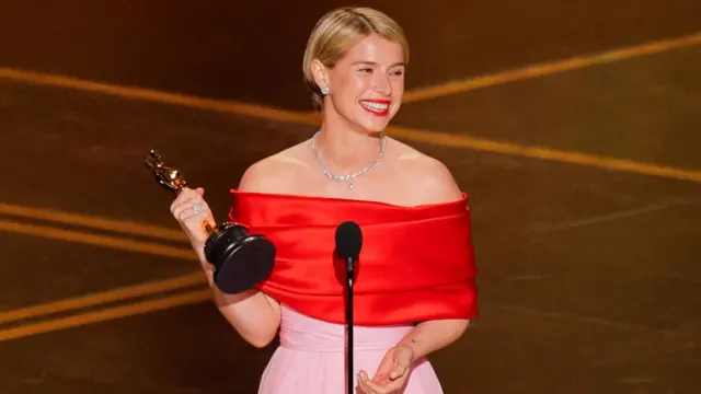 Jessie Buckley, with blonde hair styled short, and wearing a red and pink off-the-shoulder gown, grins as she holds her trophy.