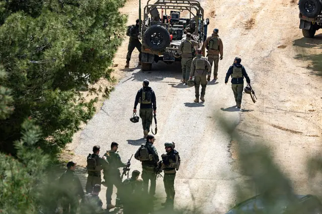 Soldiers in uniform are seen walking along a dusty road, green trees are sprinkled on the sides of them. Some of the soldiers hold weapons and others hold their helmets in their hands.