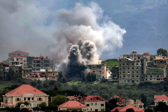 Homes with red roofs and tan exteriors are seen in the foreground of the image as smoke plumes rise in the background and a building appears to be detroyed