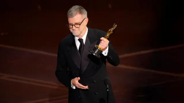 Paul Thomas Anderson, with short, greying hair, a short white beard and black-rimmed glasses, wears a black suit and tie and holds his trophy up with his left hand.