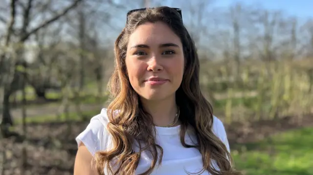Charlotte in a white t-shirt. She has brown hair and wears sunglasses on her head. She stands outside.