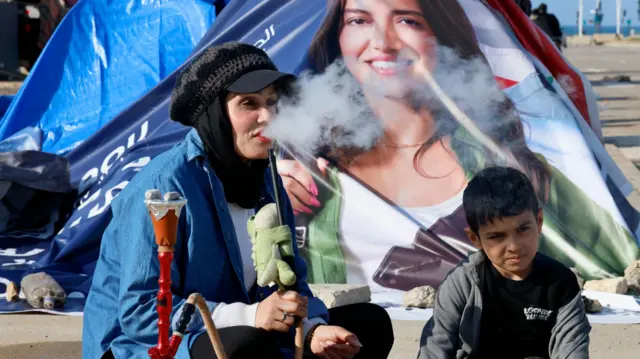 An internally displaced woman smokes a shisha waterpipe as she sits on the floor with her son outside a tent in Beirut, Lebanon, 16 March 2026.