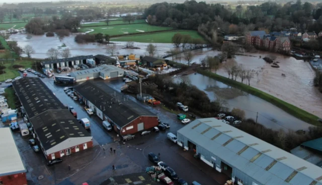 The River Otter seen from above, via a drone, overflowing onto neighbouring fields, close to industrial units in the foreground and homes in the distance.