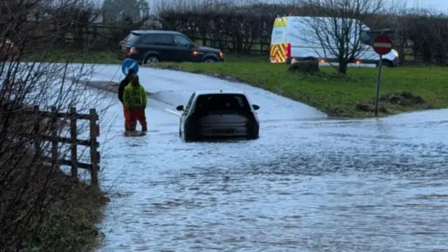 A car stuck in floodwater at Ottery St Mary. Two men are standing to the right of the car and there is a recovery truck in the background.