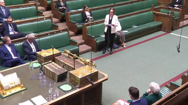 Wide high shot of House of Commons where you can see part of benches with sparse amount of MPs sitting on them and Alison Bennett standing to speak