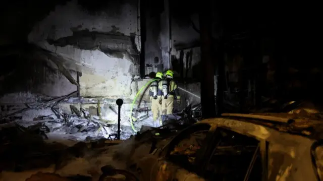 Two emergency workers hold a hose over a heavily damaged structure.