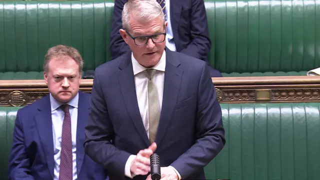 Stuart Andrew in front of the green benches in the House of Commons speaking to MPs. There are two MPs in suits sitting behind him