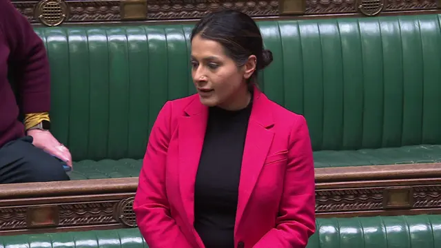 Naushabah Khan MP in a bright dark pink blazer and a high black top standing in the House of Commons in front of green benches and speaking with her hands clasped out of frame