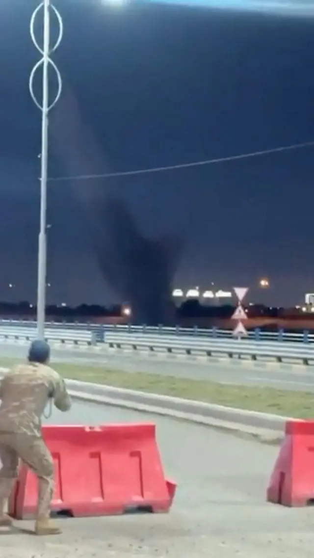 A man slightly crouches behind a red road blocker as smoke risis in the middle distance