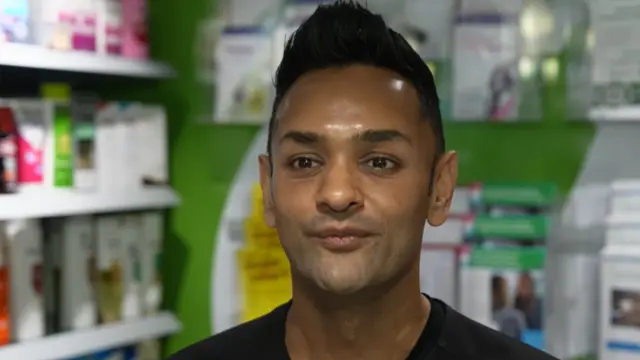 A man with short black hair wearing a black t-shirt. He is looking off to the left of the camera and is stood in a pharmacy, with prescriptions behind him
