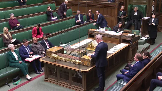 Wide shot of the House of Commons from a diagonal angle behind the opposition benches. Stuart Andrew is standing up ready to speak in front of the benches in response to the government minister speech