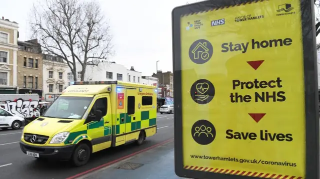 an ambulance drives past a poster saying 'stay home, protect the nhs, save lives'