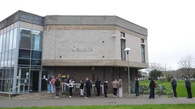 students queue on the pavement in front of a kent university building in canterbury. some are wearing masks. many are social distancing.