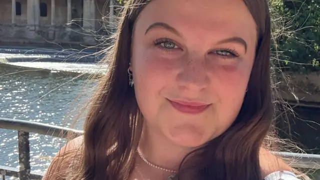 Ella stands in front of a river. she has long brown hair and wears a white top with black dots.