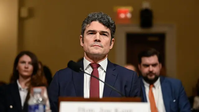 Joseph Kent sits at a desk in front of the camera. Others sit behind him and have been blurred in the image