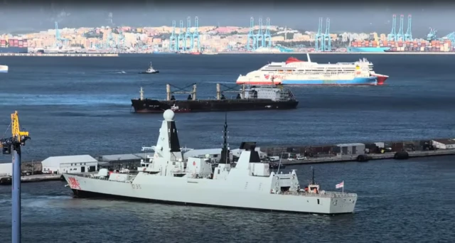 HMS Dragon, a grey warship, seen at Gibraltar while in the background a ferry and dockside cranes can be seen on the opposite side of the bay