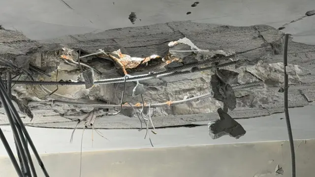 A large hole in the ceiling of he apartment ringed with broken concrete and twisted metal rods