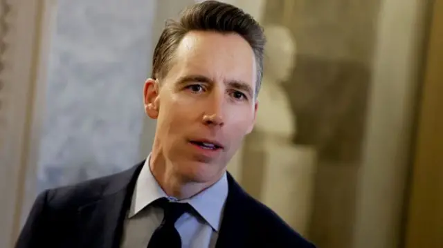 Republican Senator Josh Hawley in a dark suit against a beige background.