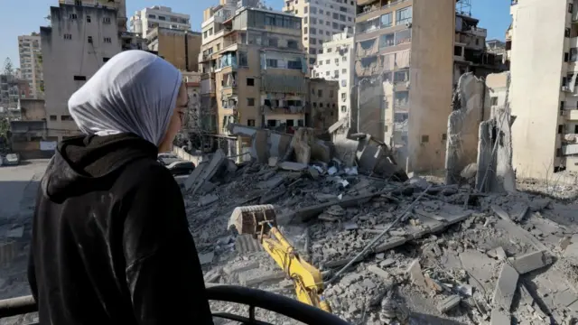 Woman wearing a grey hijab and black hoodie has her back to the camera and looks out over a pile of rubble and a digger going through it, which was a building destroyed in strikes