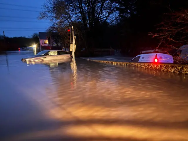 Two vehicles - a 4x4 and a white van - are submerged in flood water along a road. A house is in the distance.