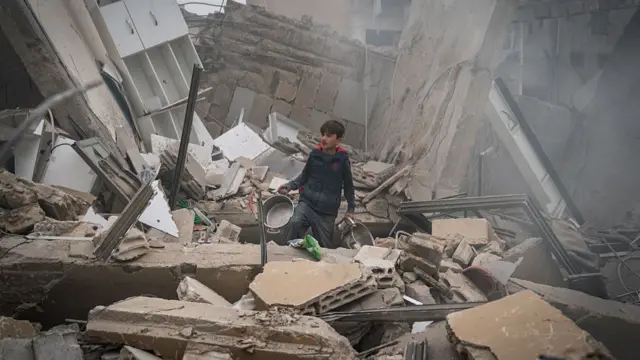 a young boy holding a kitchen pot as he stands amid a mountain of rubble