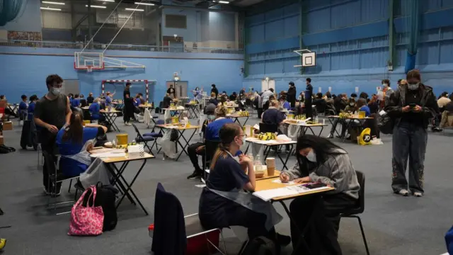 Students and nurses sit at individual desks spread out across a sports hall as vaccines continue to be rolled out