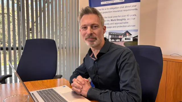 Mortgage advisor Mark Doughty sitting at a wooden desk with a laptop in front of him. He's looking to his left towards the camera, his hands in front of him with his fingers intertwined. He's in a dark blue shirt, sitting on a black cushioned chair