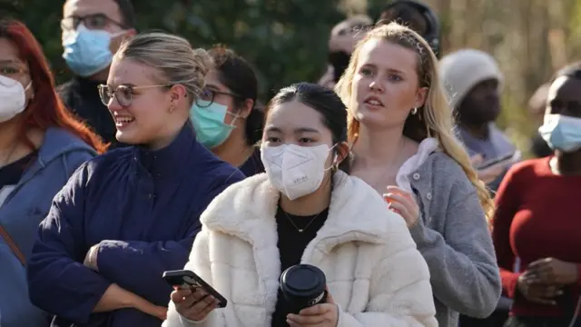 Several women are seeing in a queue for meningitis vaccinations at a vaccine centre. The woman in the centre of the image is wearing a PPE face mask and is wearing a fluffy white jacket while holding her phone and a coffee cup.