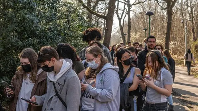 Dozens of students queue for the Meningitis B vaccine on a path lined with trees in the sun