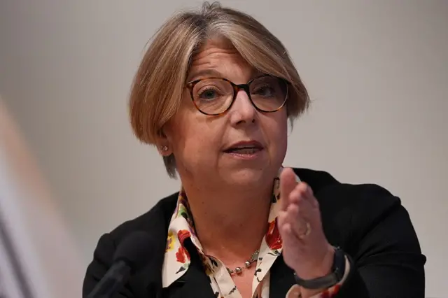 Sarah Breeden (woman in her 50s, short blonde hair) mid-speech. She's gesturing with her left hand, wearing a black blazer and floral shirt