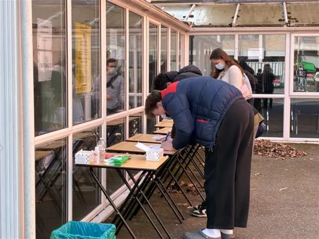Two people lean over tables outside, while another stands behind them wearing a facemask