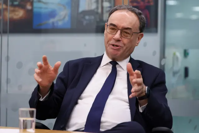 BoE governor Andrew Bailey mid-speech whilst sitting on a chair at a wooden table wearing a dark blue suit
